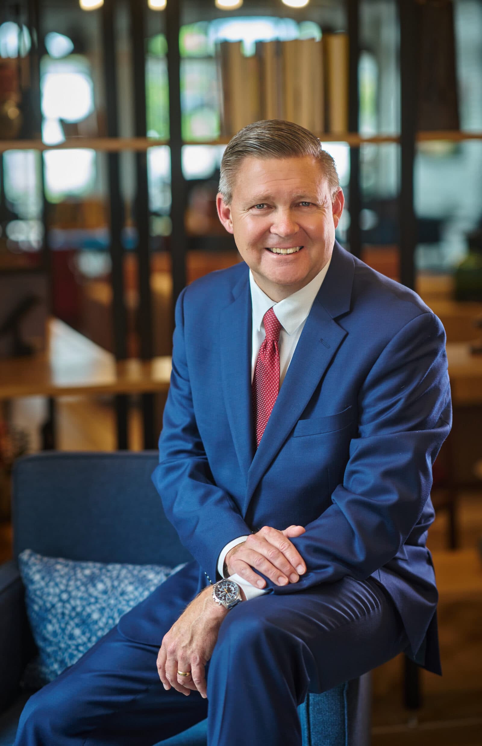 David is a savvy looking middle aged white man in a navy blue suit, red tie with white dots. He sits on a chair and smiles at you.