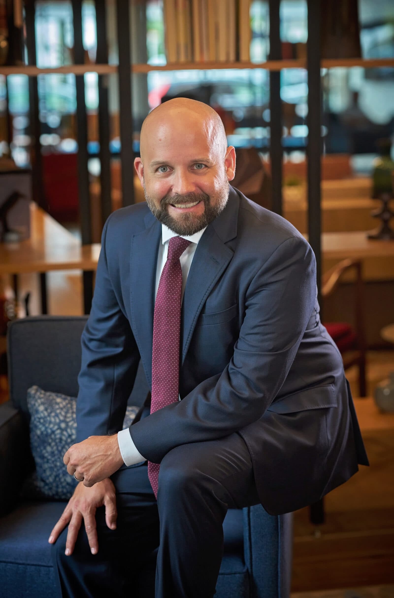 Peter is a bearded, bald man wearing a suit and red tie with white dotes. He sits and smiles at you.