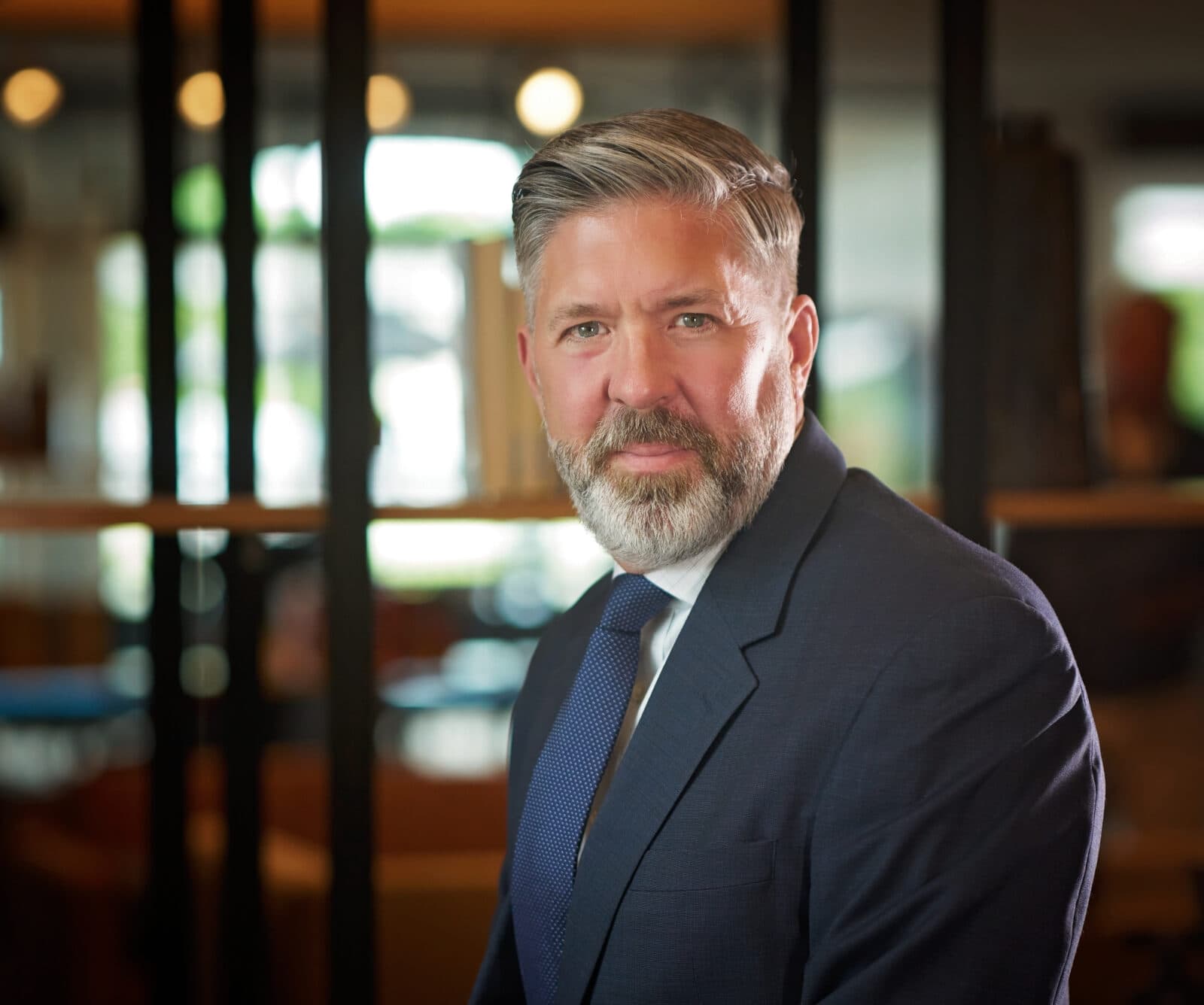 Trawn is a distinguished looking gentleman with a graying beard and blond / gray hair. He wears a suit with blue tie, and smiles at you.