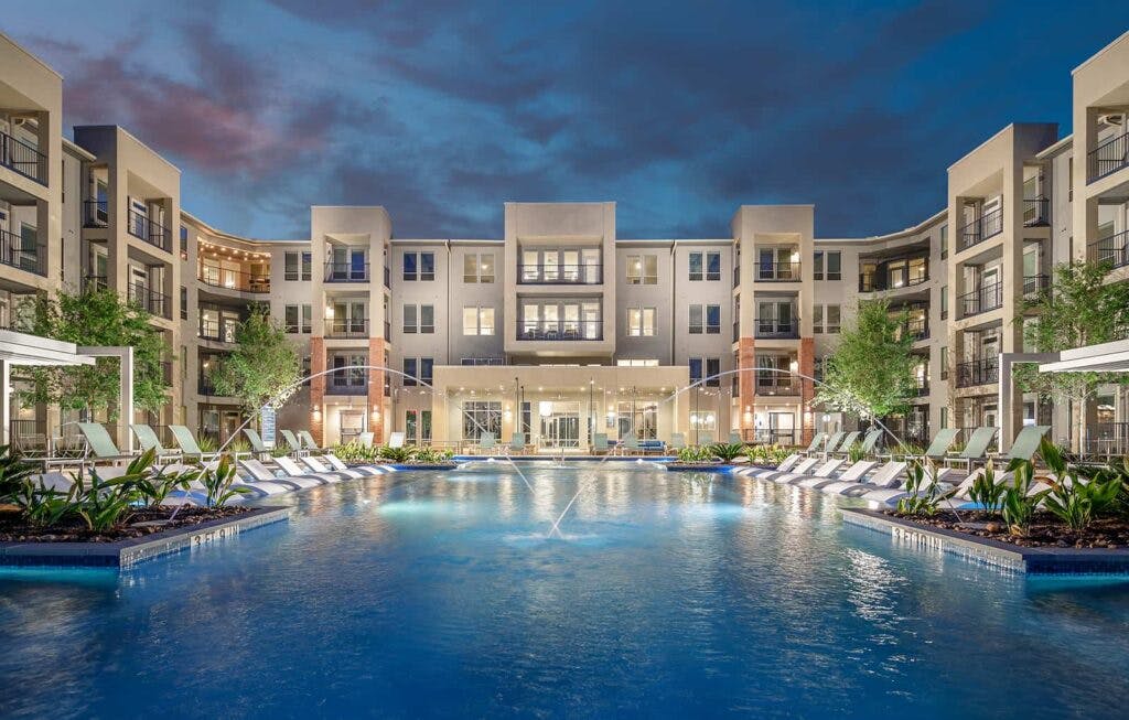 A square, blue, and inviting pool rest languidly in the middle of a classy apartment complex. Twin fountains arc streams of water into the pool from either side.