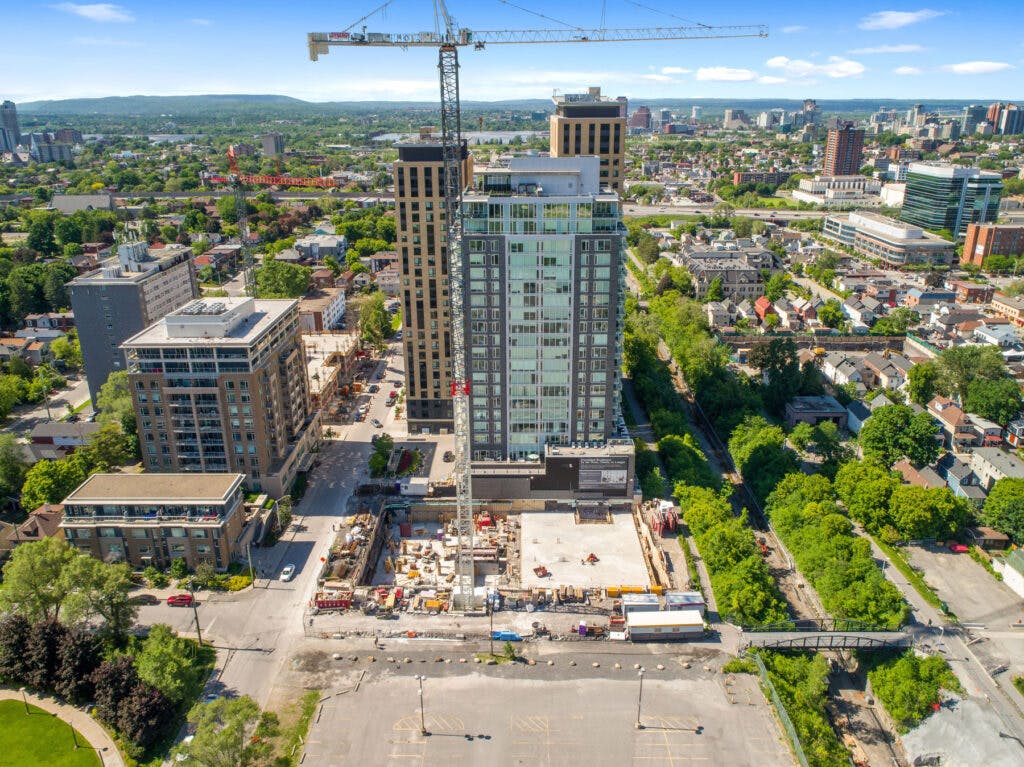 An aerial view of a construction site in a city. It is surrounded by trees and buildings. The construction site is a hive of activity, with people and machinery working together.