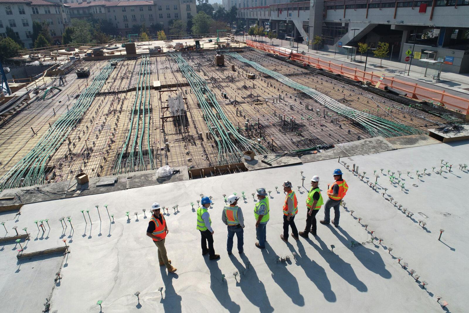 A group of construction workers standing on top of a construction site. The workers are wearing hard hats and safety vests, and they are looking down at the construction site.