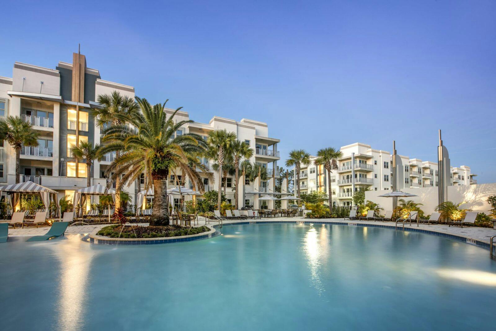 Exclusive apartment buildings stands before a glistening swimming pool surrounded by palm trees and bushes.
